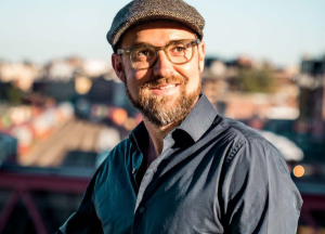 Man with a beard, mustache and dark rimmed classes in a long sleeve blue button down shirt and a brown checked newsboy cap, smiling and looking just to the right of the camera.