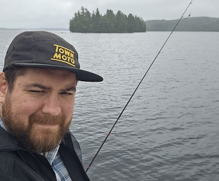 Man with a beard and mustache wearing a blue checkered shirt and light black rain jacket. He's wearing a black ball cap that says 'Town Moto' in yellow text. He's holding a fishing rod and is on a boat in a lake, with trees in the background on a foggy day.