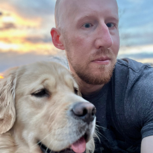 Man with sandy beard/mustache and no hair in a grey shirt looking at the camera. He's posing with a gold retriever dog with its tongue slightly out. There's a sunset in the background dappled by grey clouds.