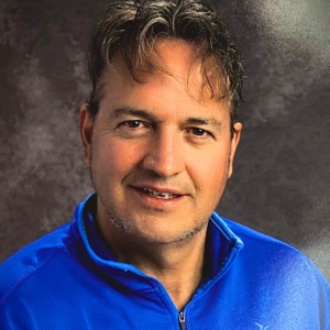 Man posing for a photo against a grey background. He is wearing a blue athletic zip-up shirt and has hair that's short on the sides and longer over the forehead.