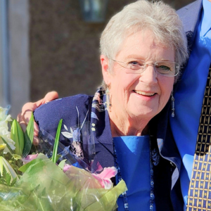 Older woman with short grey hair in a blue suit, holding flowers. She's wearing glasses and smiling at the camera, leaning against a person in a suit who has been cropped out of the photo.