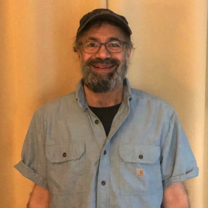 Man with salt-and-pepper beard, ball cap, and dark-rimmed glasses. He's standing and smiling into the camera. He's wearing a dark undershirt and blue short-sleeved button up.