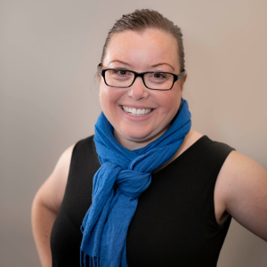 Woman with glasses, hair pulled back, and a blue scarf with hands on hips, smiling into camera