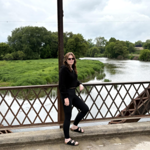 woman in all black leaning against a diamond-patterned fence and pole, with a river and green mossy land and trees in the background