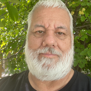 Selfie of man with white hair and beard, with a background of green leaves. He's wearing a black T-shirt