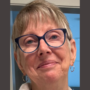 Close up of an older Caucasian woman with very short blondish hair, blue glasses and gold hoop earrings.