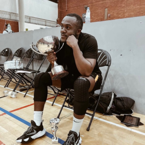 Tall Black man in black shirt, shorts, and white socks with basketball sneakers, sitting in a fold up chair on a basketball court. He is leaning forward, one elbow on his knee and is holding a trophy.