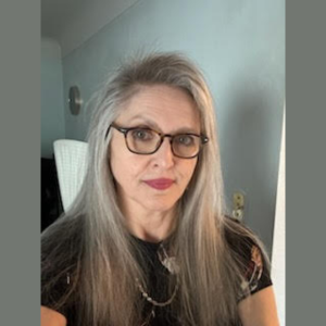 Caucasian woman with long grey blonde hair and glasses in a black patterned short-sleeve shirt, looking at the camera.